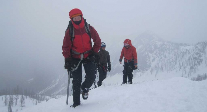 Three people in mountaineering gear make their way across a snowy, mountainous landscape.